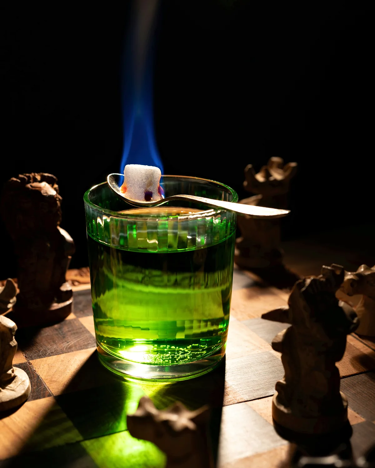 A glass of green absinthe with a flaming sugar cube on a dark chessboard