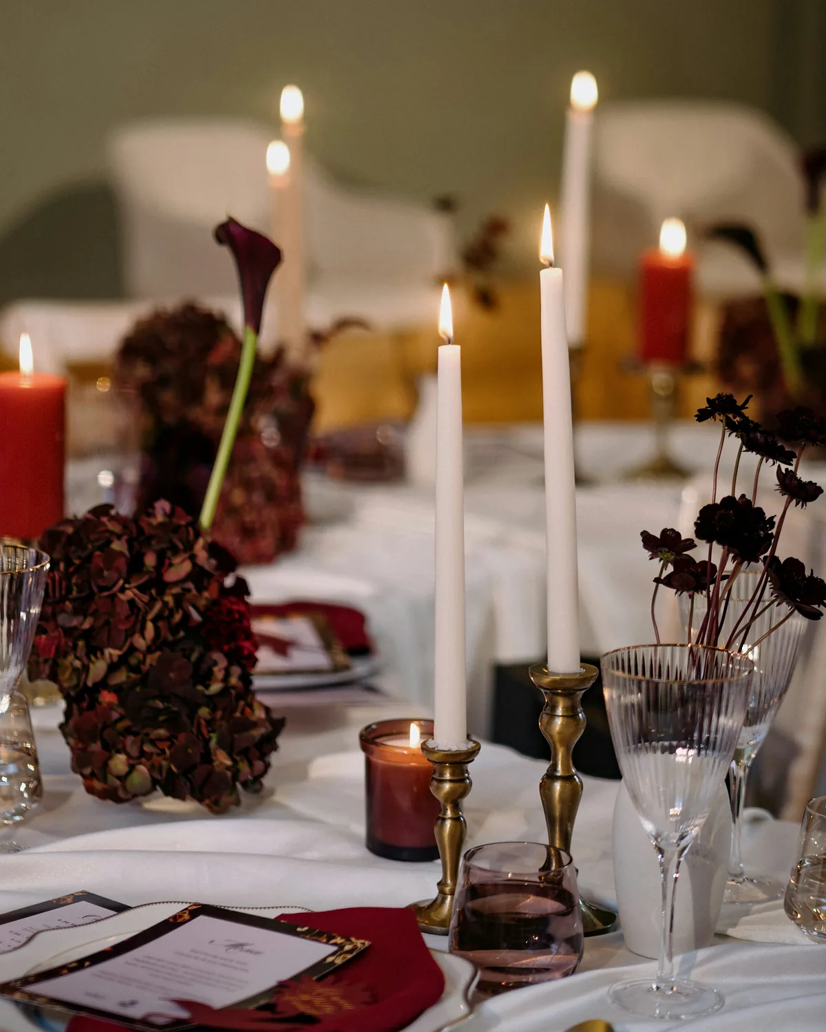 A candlelit dinner table set with white tapers, brass candlesticks and dark burgundy florals
