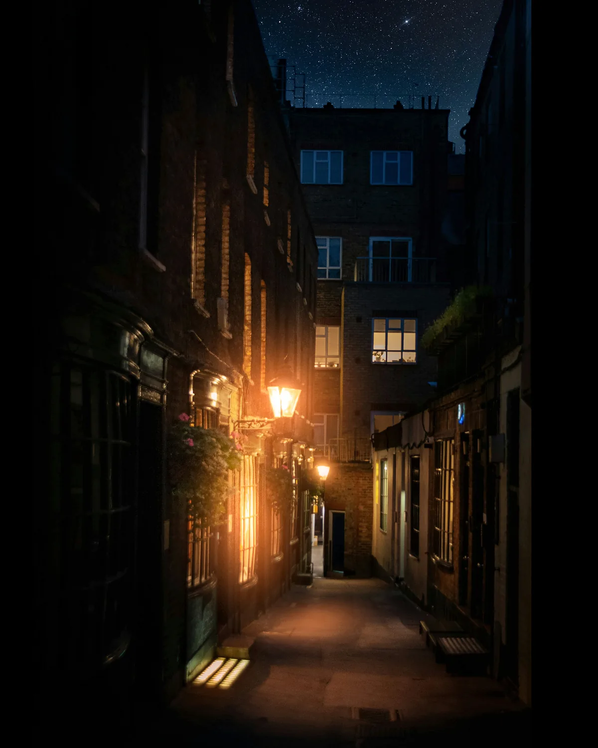 A narrow London backstreet at night, lit by a single old lamp between dark brick buildings