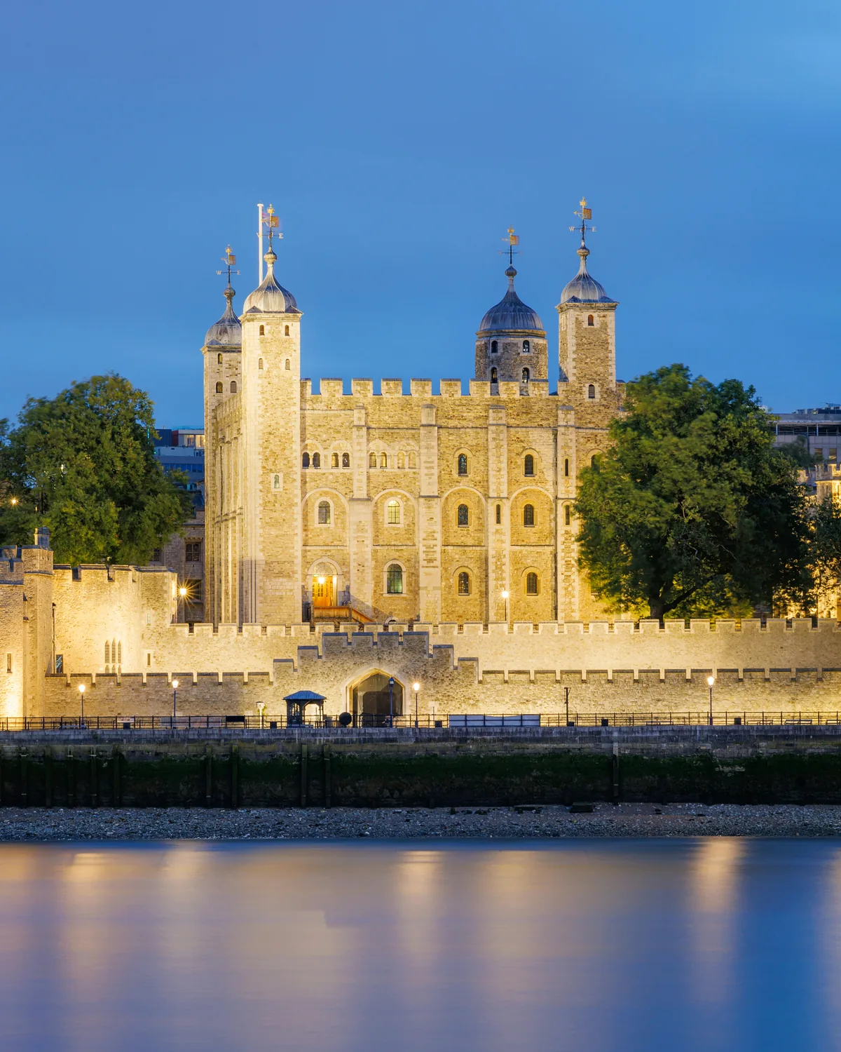 The floodlit White Tower and walls of the Tower of London at blue hour, reflected in the Thames