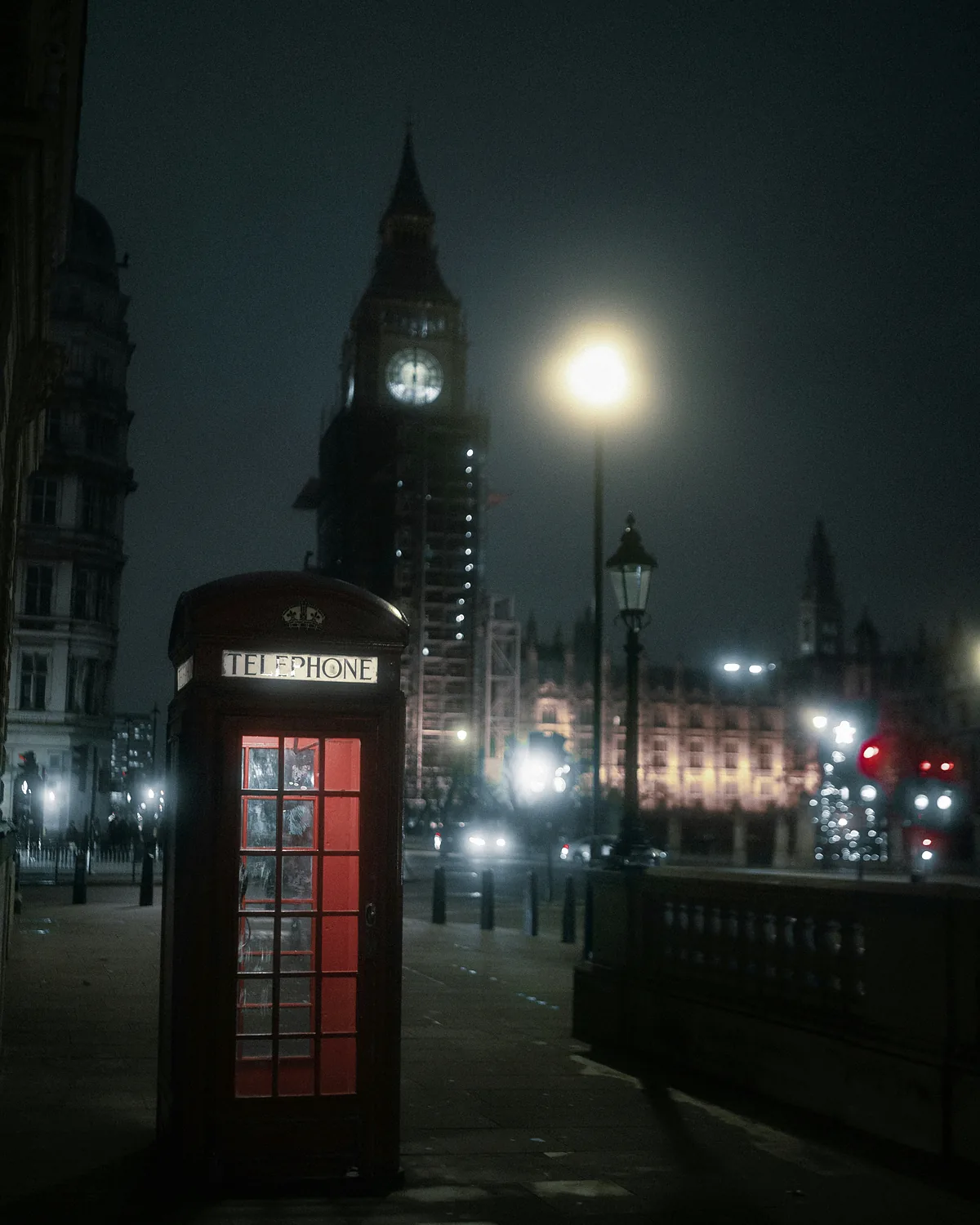 A red telephone box glowing on a dark Westminster street, with Elizabeth Tower lit behind it
