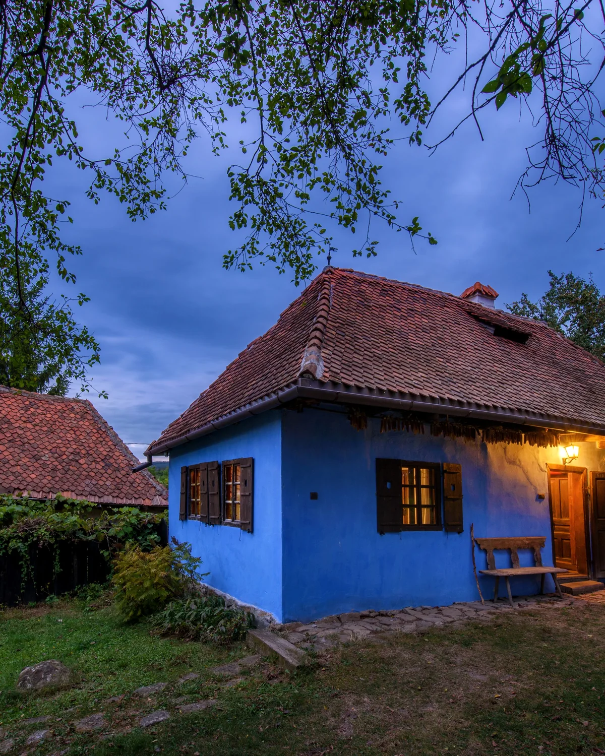 The Blue House at Count Kálnoky's, a bright blue village cottage with a red tiled roof and a lit porch at dusk