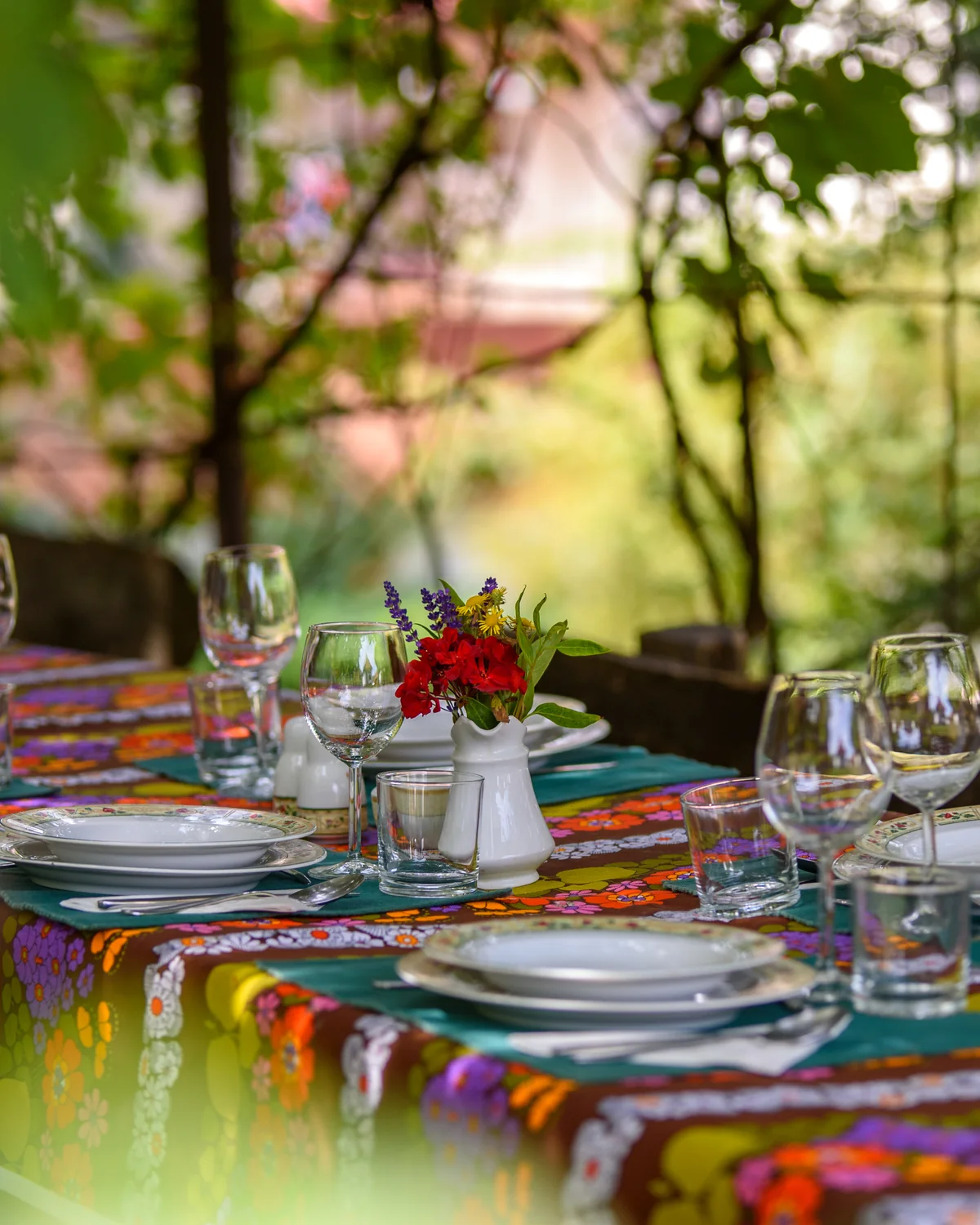 An outdoor table set with a colorful striped cloth, glasses and a small bunch of red flowers in a green garden