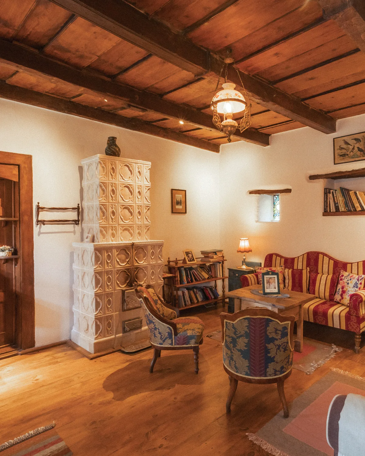 The Prince's Suite living room, with a white tiled stove, a red striped sofa, bookshelves and a beamed ceiling