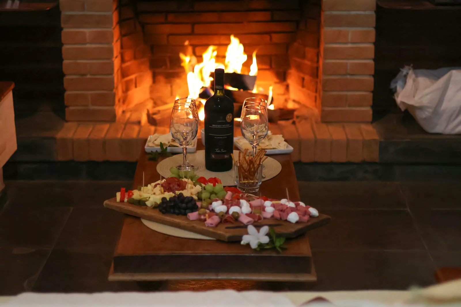 A crackling brick fireplace behind a board of charcuterie and a bottle of red wine