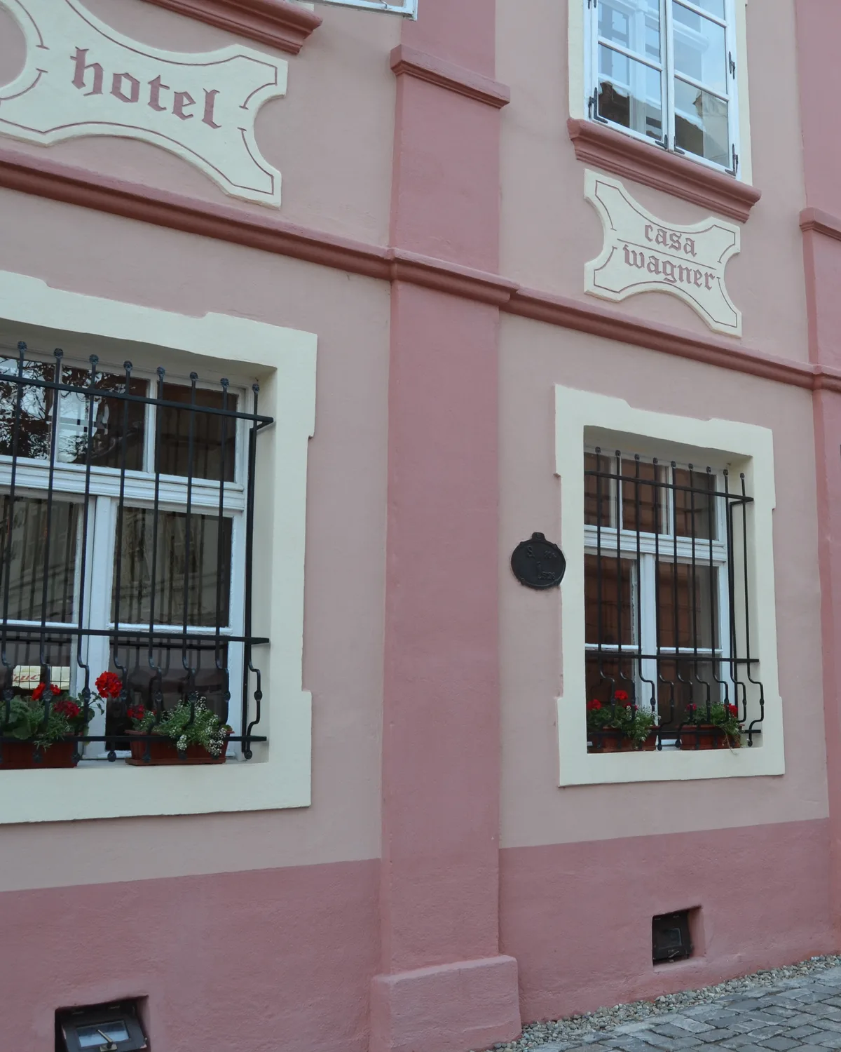The pink facade of Casa Wagner in Sighișoara, with its hotel and casa wagner plaques above barred windows and red geraniums