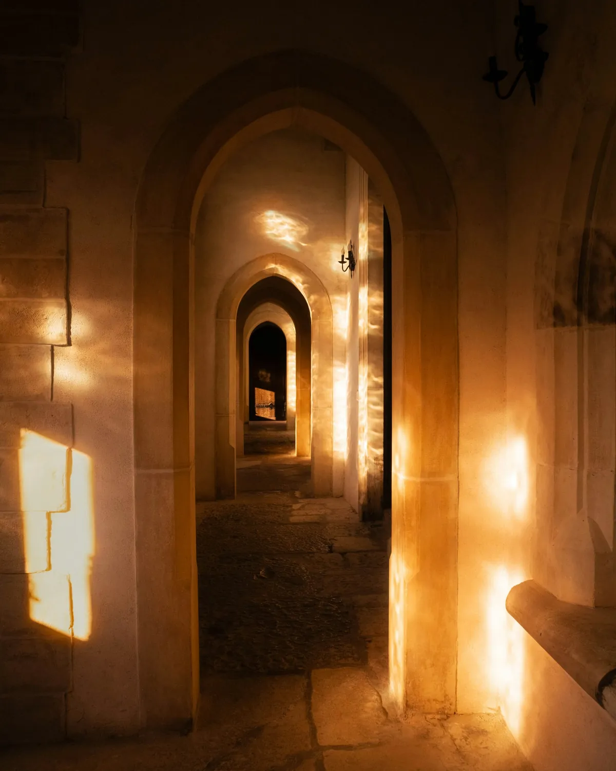 A dim stone corridor at Corvin Castle lit by warm lamps, arches receding into shadow