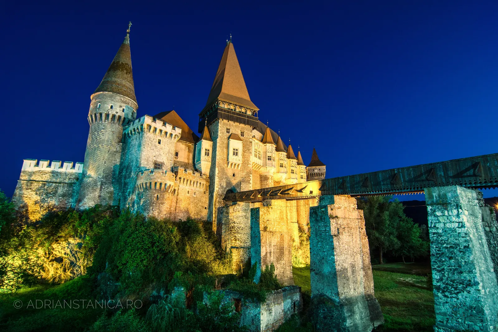 Corvin Castle lit gold at night, its pointed towers and drawbridge glowing against a deep blue sky