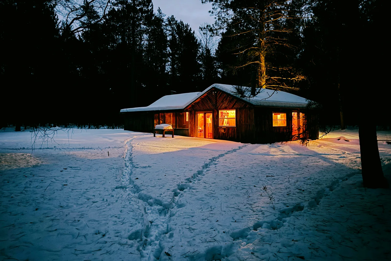 A wooden cabin in the snowy woods at dusk, its windows glowing warm and footpaths leading to the door