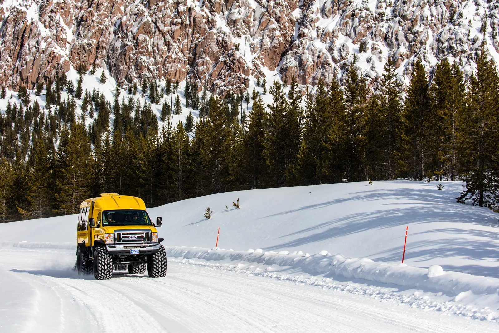 A yellow snowcoach on a groomed snowy road below a frosted cliff and pine forest in Yellowstone