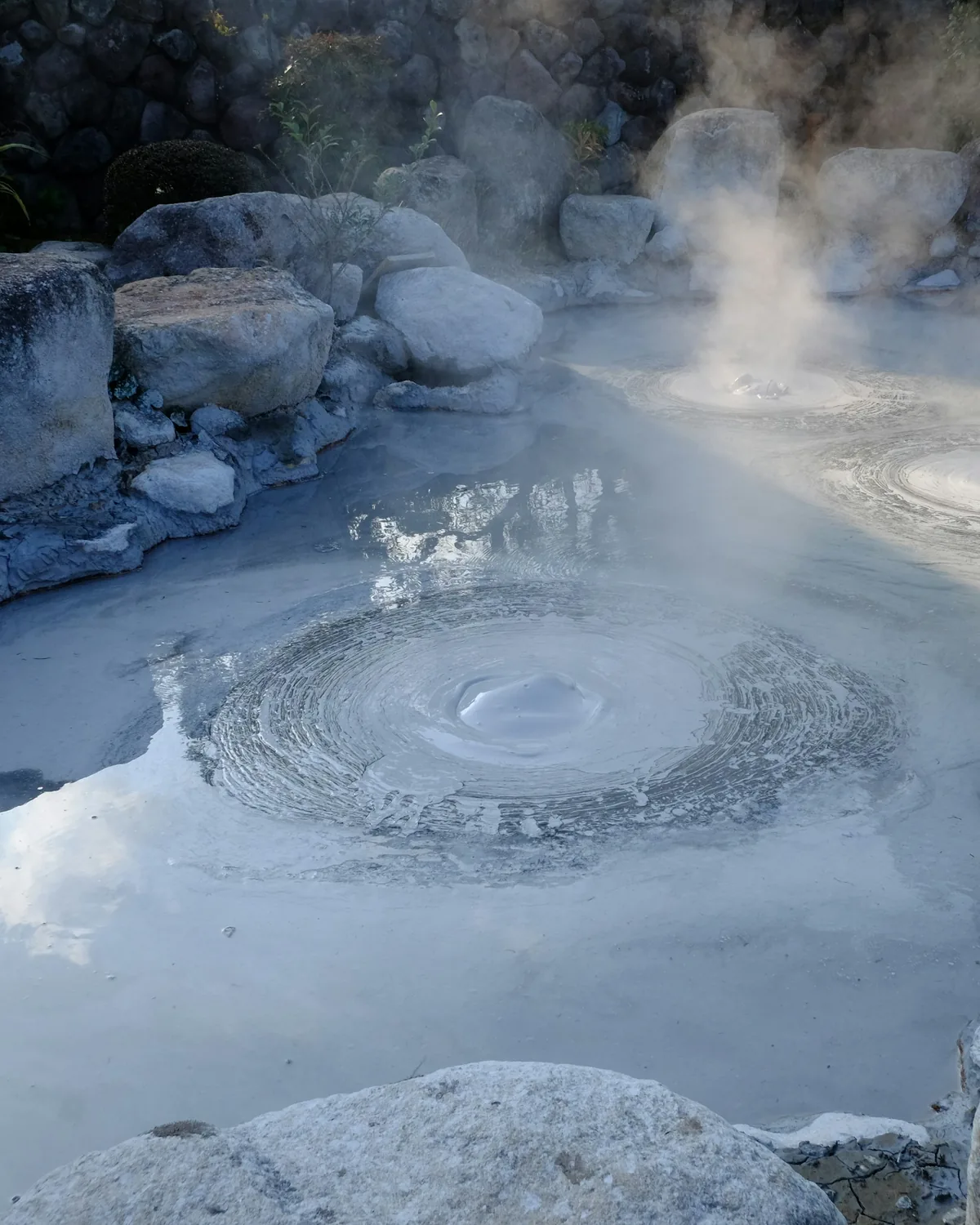 Steam curling off a natural hot spring pool ringed with smooth rocks on a cold day