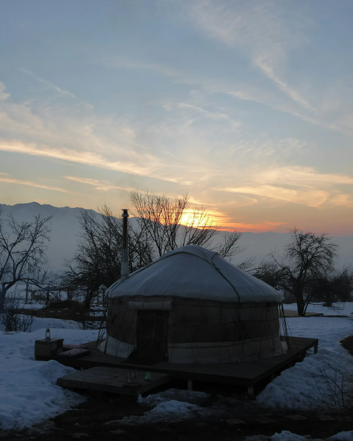 A round yurt in fresh snow under a glowing sunset sky with bare trees and distant hills