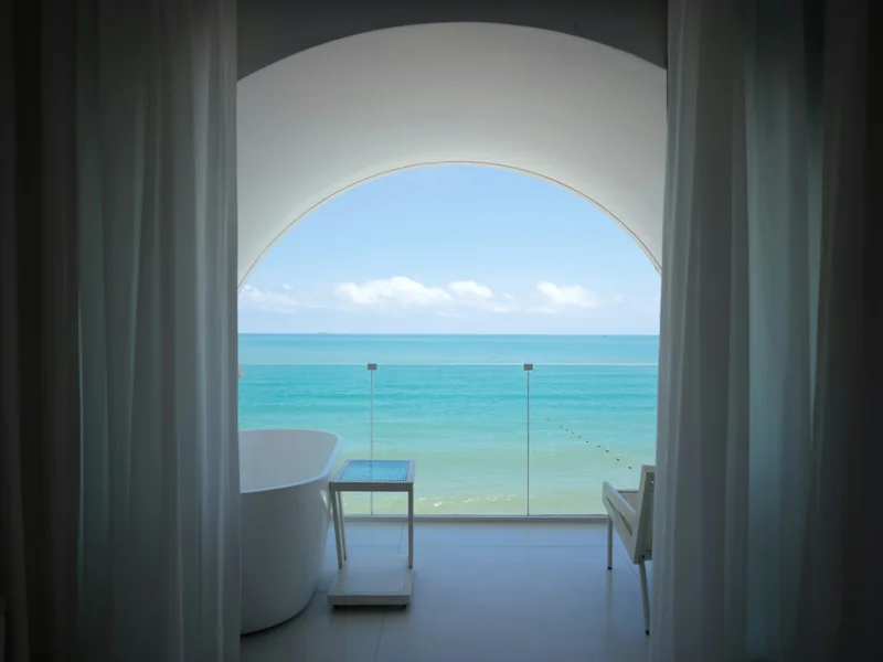 A sunlit hotel bathroom with a freestanding tub, sheer curtains and an arched window open to a turquoise sea