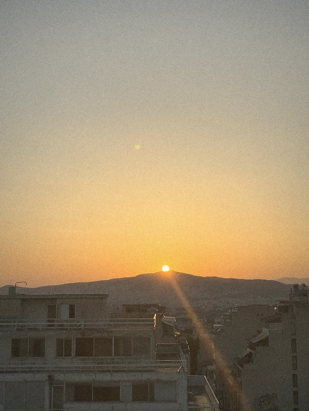 The sun setting behind a hill over the rooftops of Athens in an orange sky