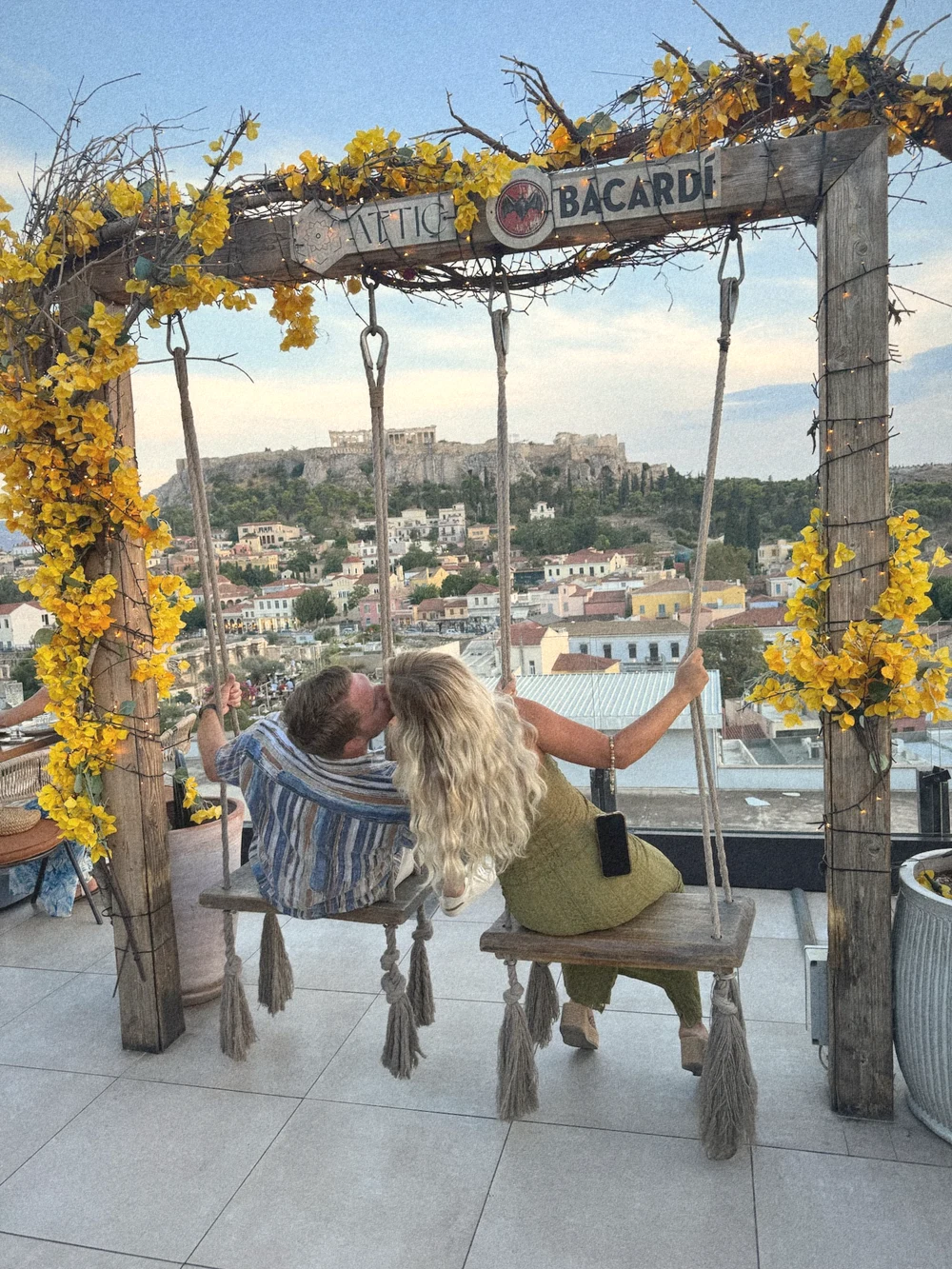 The two of us kissing on a flower-covered rooftop swing at Attic, with the Acropolis behind us