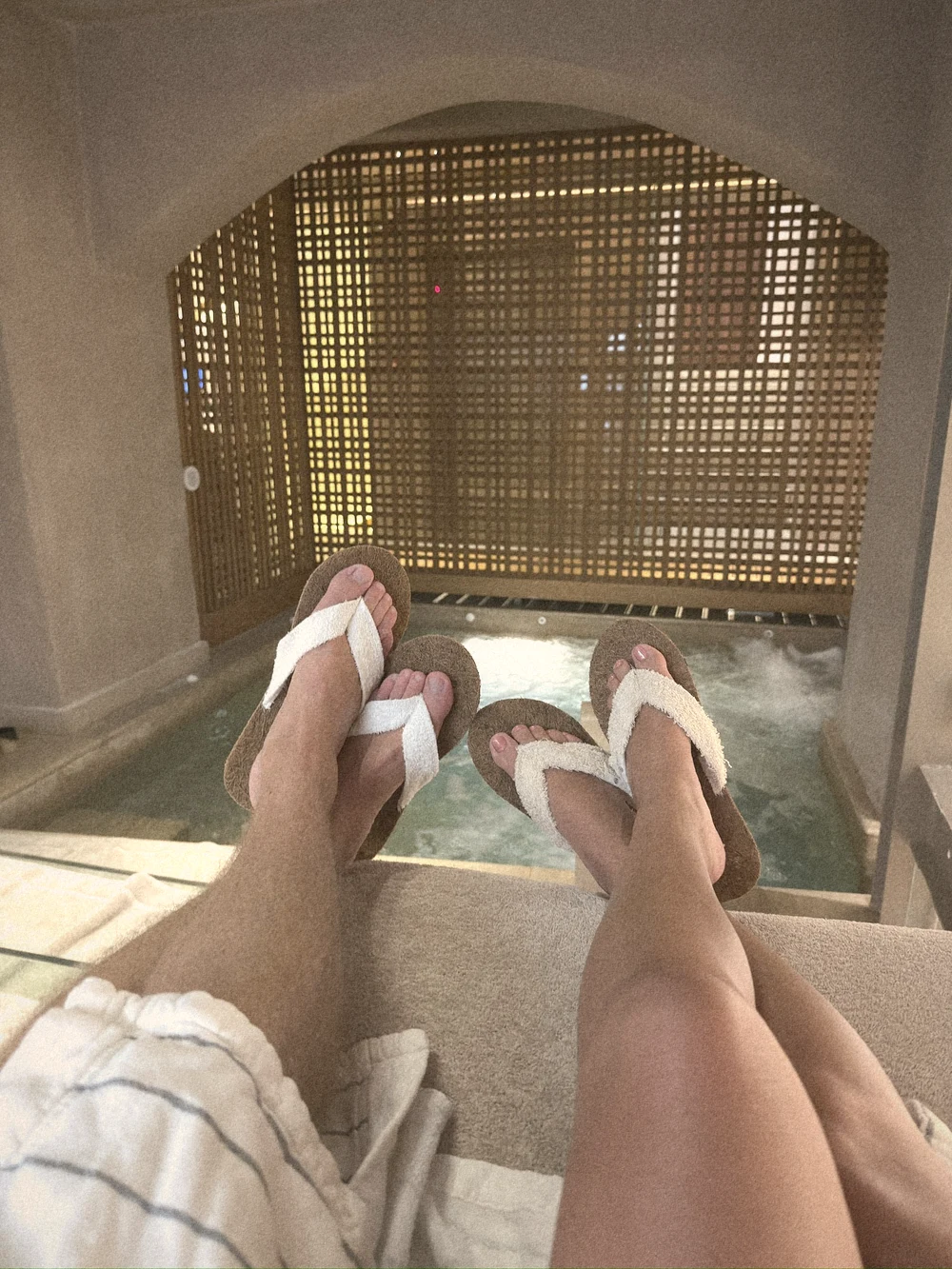 Two pairs of feet in spa sandals in front of a softly lit hydro-massage pool
