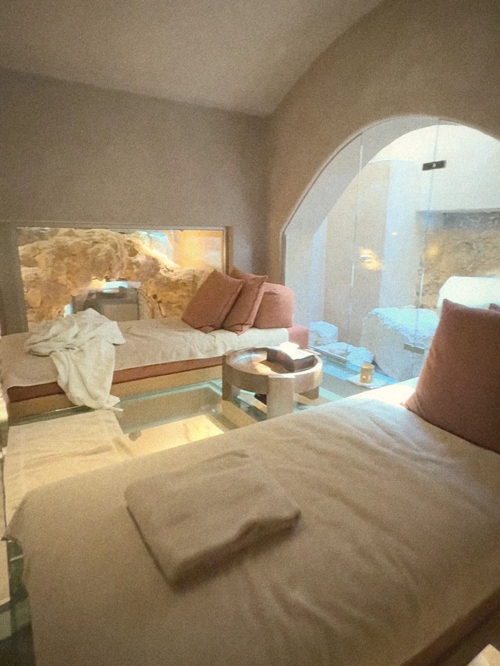 A calm, softly lit spa room with daybeds beside ancient stones behind glass
