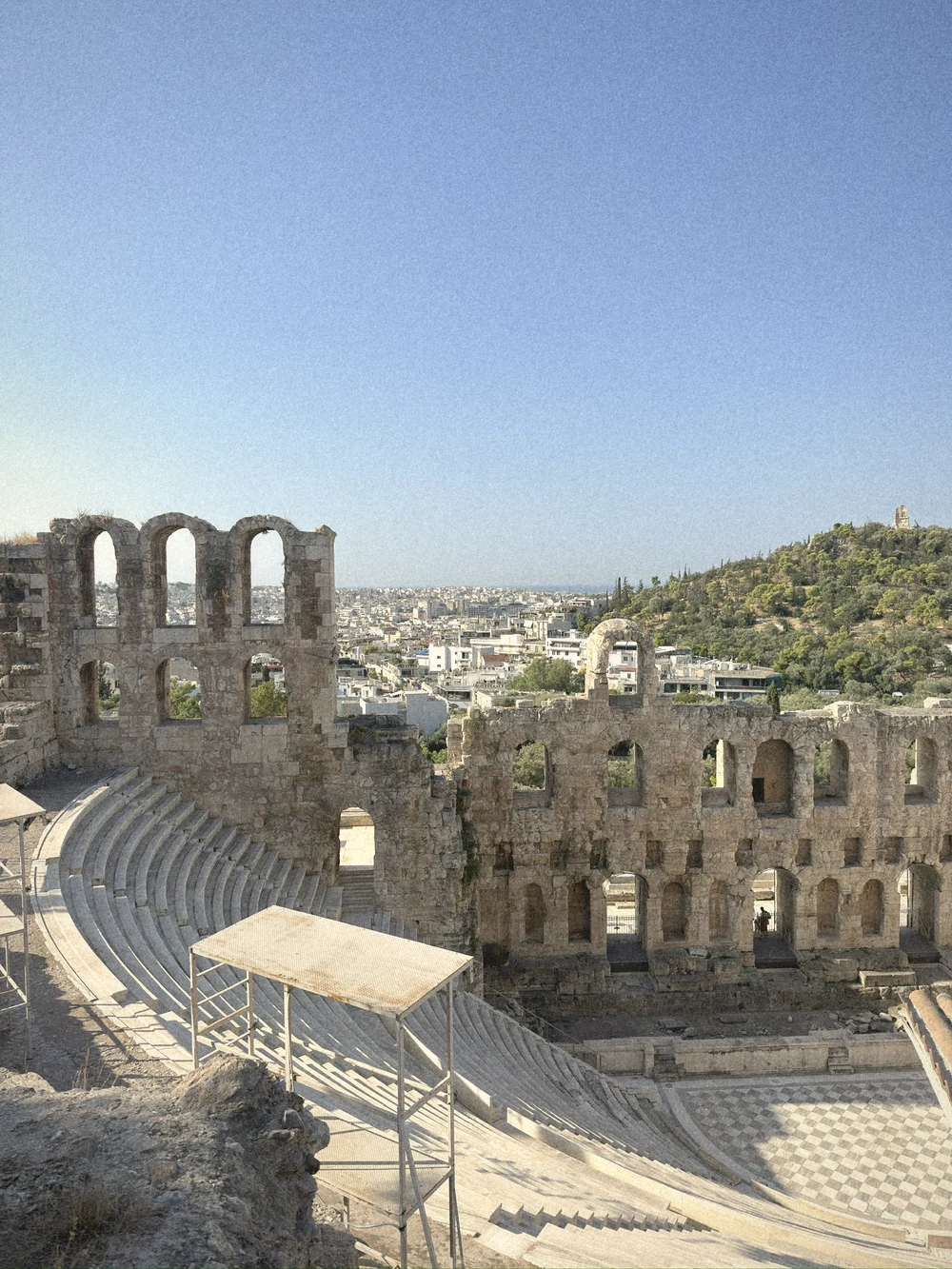 The arched stone walls and marble seating of the Odeon of Herodes Atticus, with the city and a green hill beyond