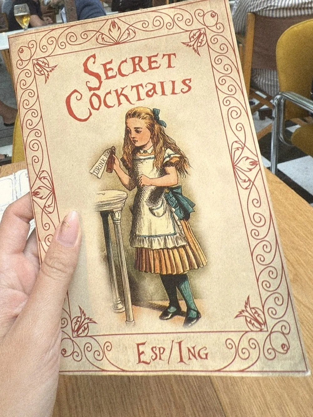 A hand holding the Secret Cocktails menu, illustrated with Alice in Wonderland, on a garden terrace table