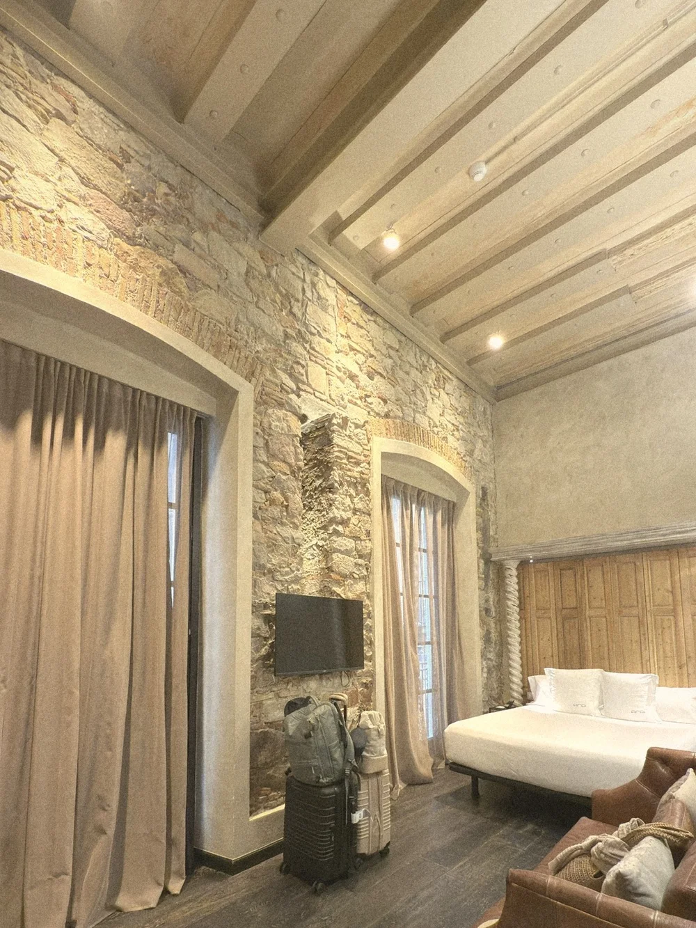 A room at Arai Aparthotel: an exposed stone wall, whitewashed beamed ceiling, tall linen curtains, a white bed against a carved wood wall, and two carry-on suitcases side by side
