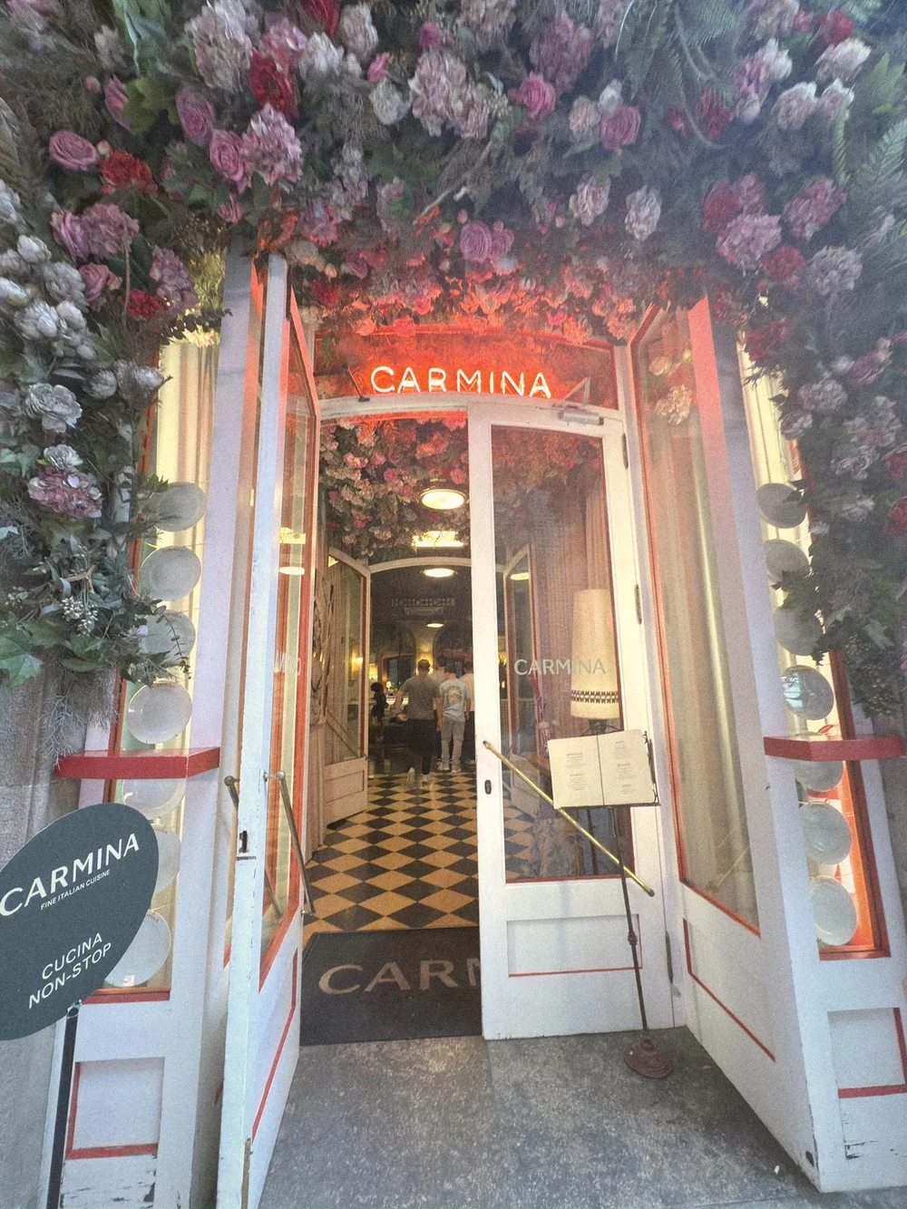 Carmina's entrance under a canopy of pink and red flowers, with a red neon CARMINA sign and a checkerboard floor