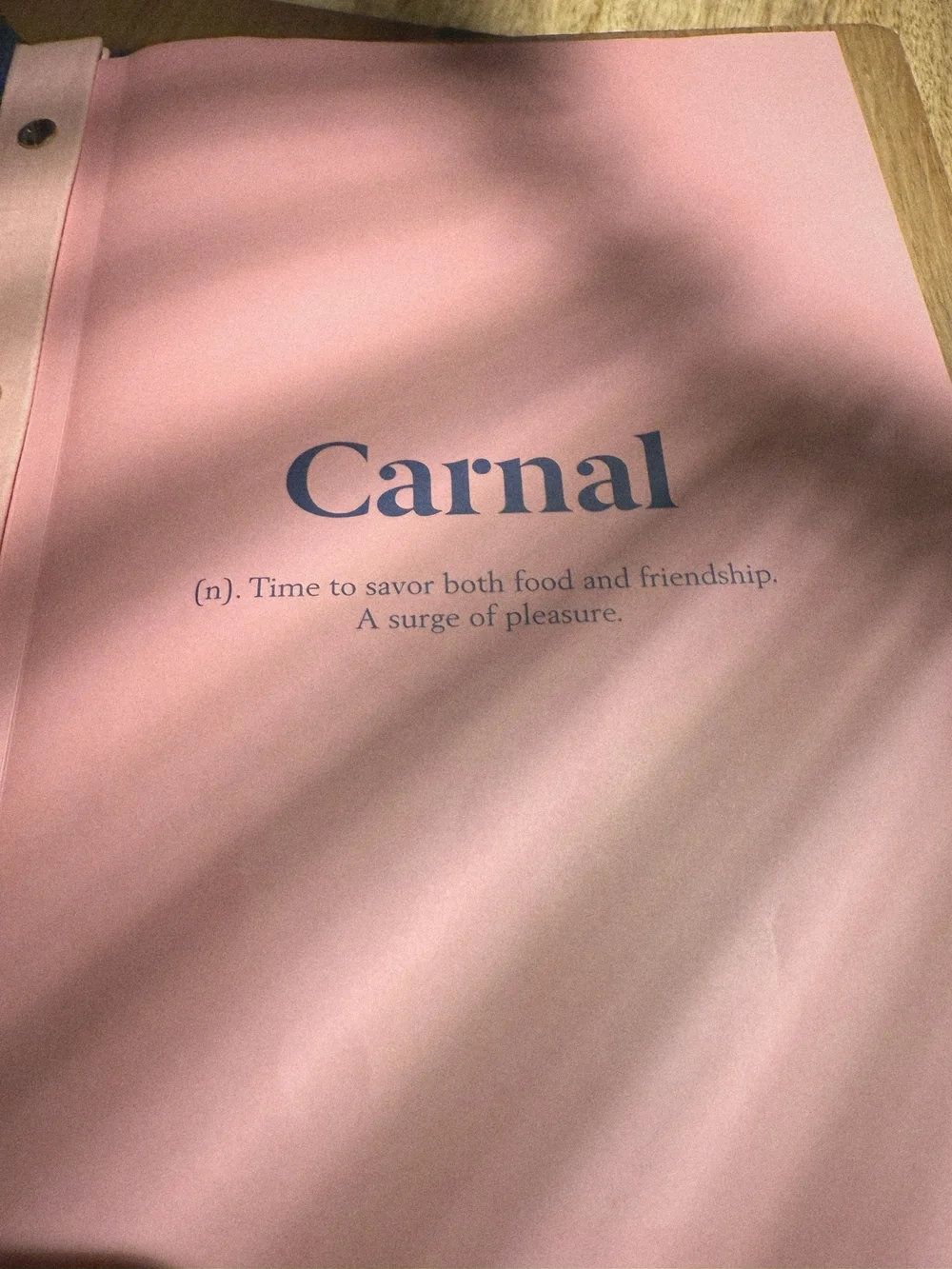 The pink Carnal menu cover in dappled light: Carnal, (n). Time to savor both food and friendship