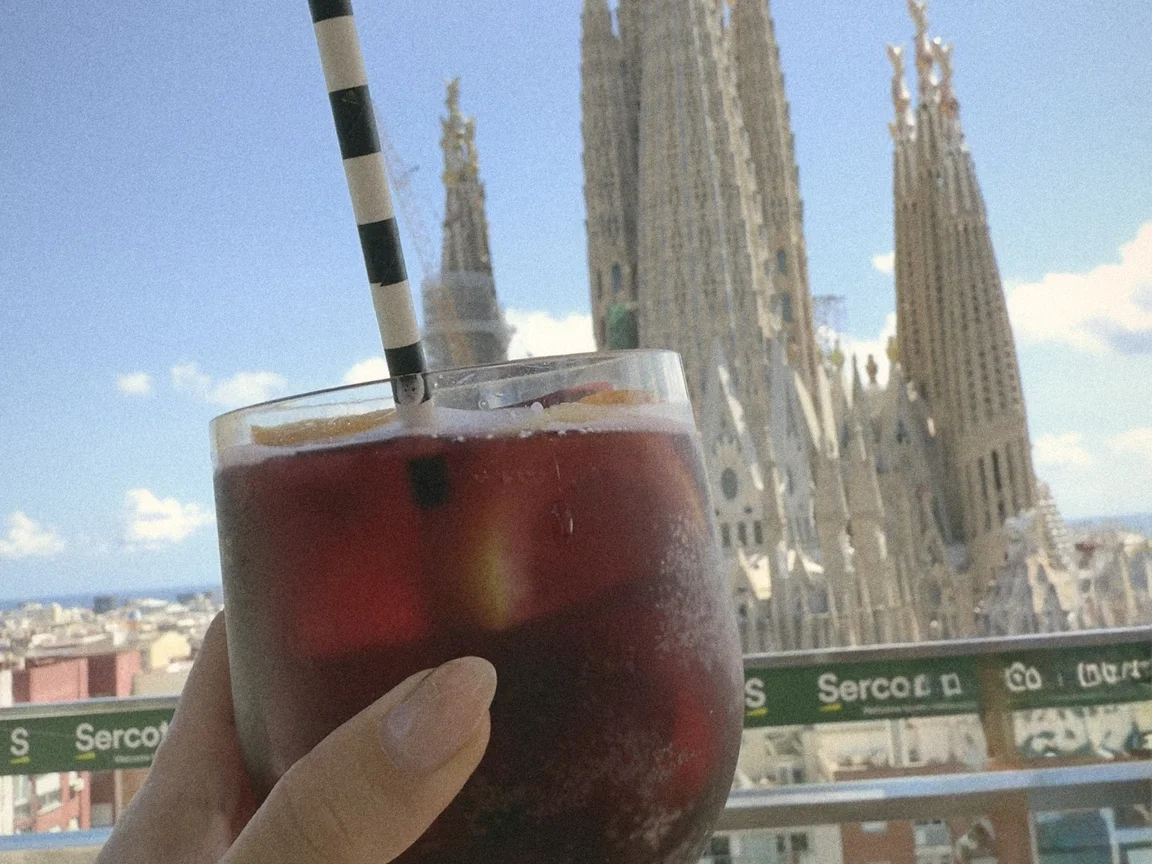 A hand holding a glass of red sangria with a striped straw on a rooftop terrace, the spires of the Sagrada Família behind it under a blue sky