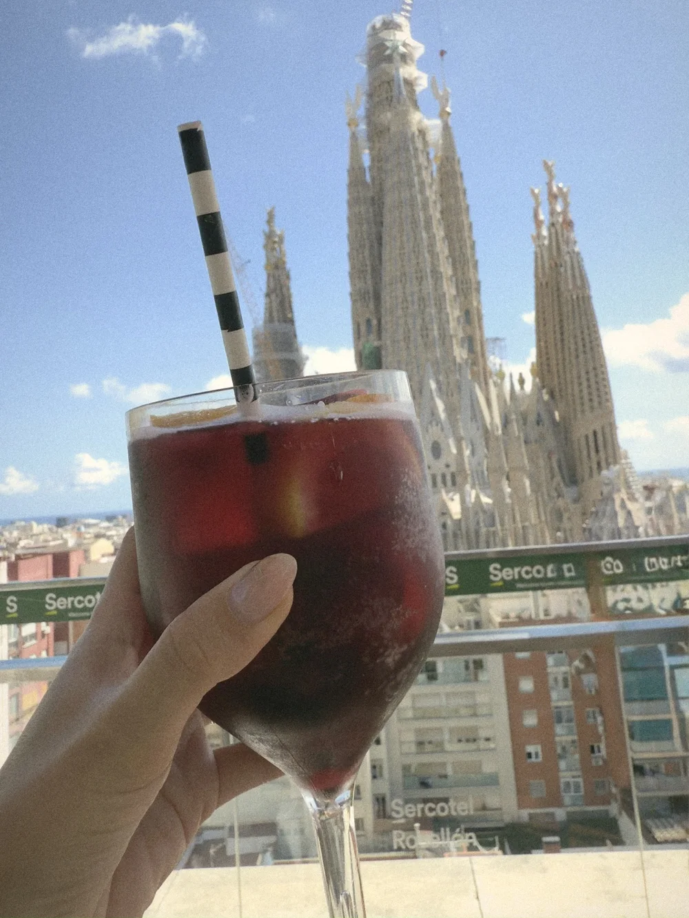 A hand holding a glass of red sangria with a striped straw on the rooftop terrace, the Sagrada Família behind it