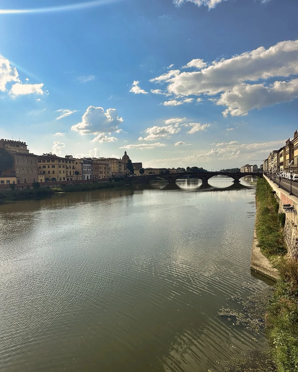 The Arno river under a blue sky with a stone bridge and riverside buildings in the distance