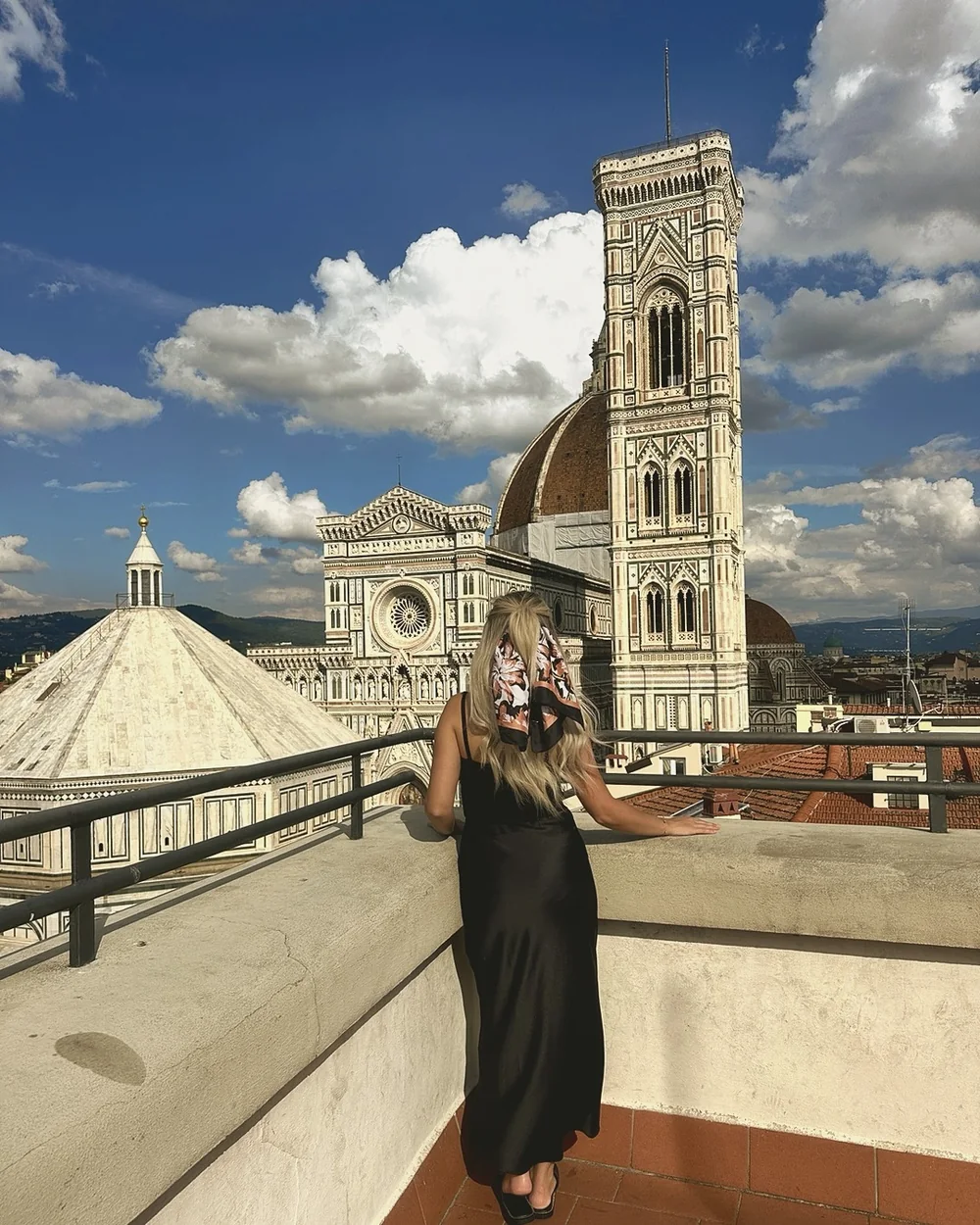 Giotto's bell tower and the cathedral seen from a rooftop terrace on Piazza del Duomo, with a woman in a black dress seen from behind