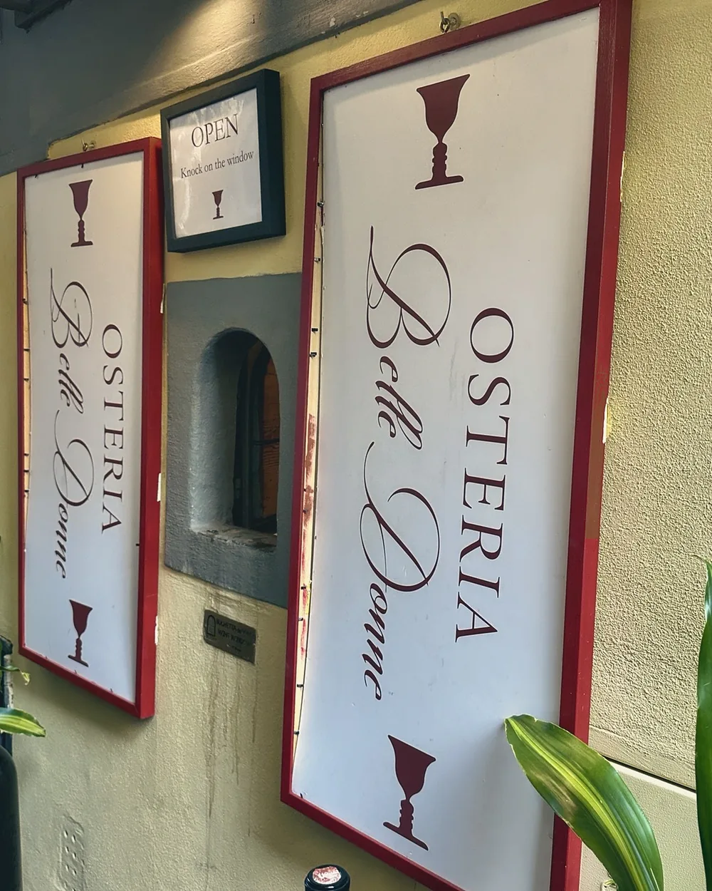 The small arched wine window between two Osteria Belle Donne signs on a Florence wall