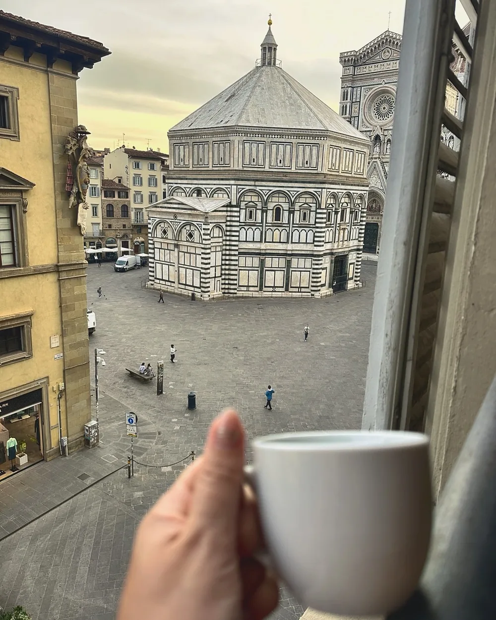 A hand holding an espresso cup at an open window above the Baptistery and Piazza San Giovanni