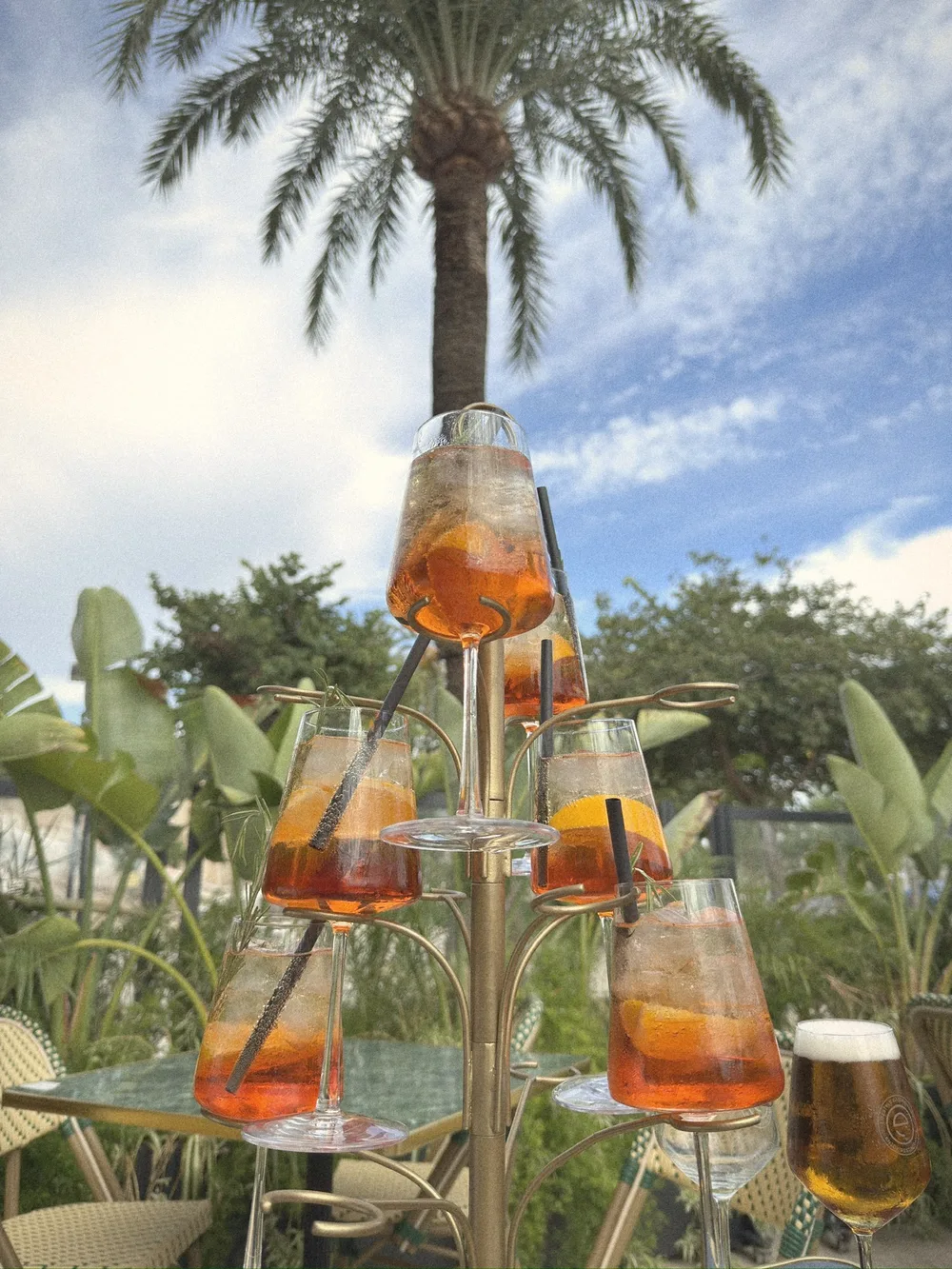 Aperol spritzes in stemmed glasses on a gold tiered drinks tree, with palms and blue sky behind