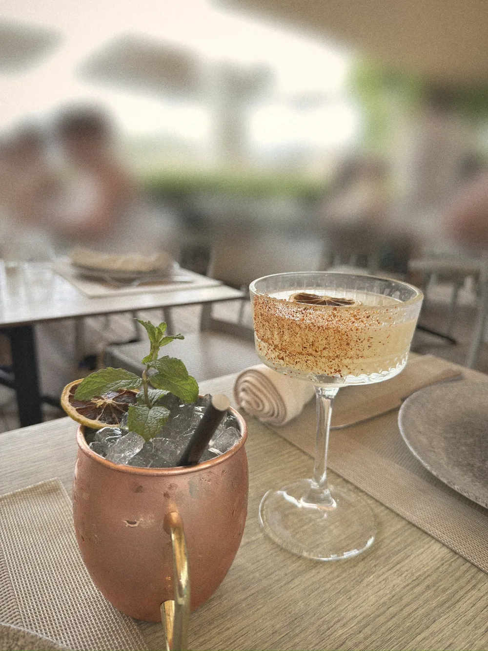 A cocktail in a copper mug with fresh mint beside a coupe with a spiced rim on a terrace table