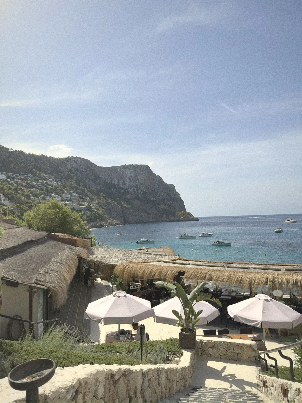 White umbrellas and thatched roofs stepping down towards a turquoise bay with boats at anchor and a high headland