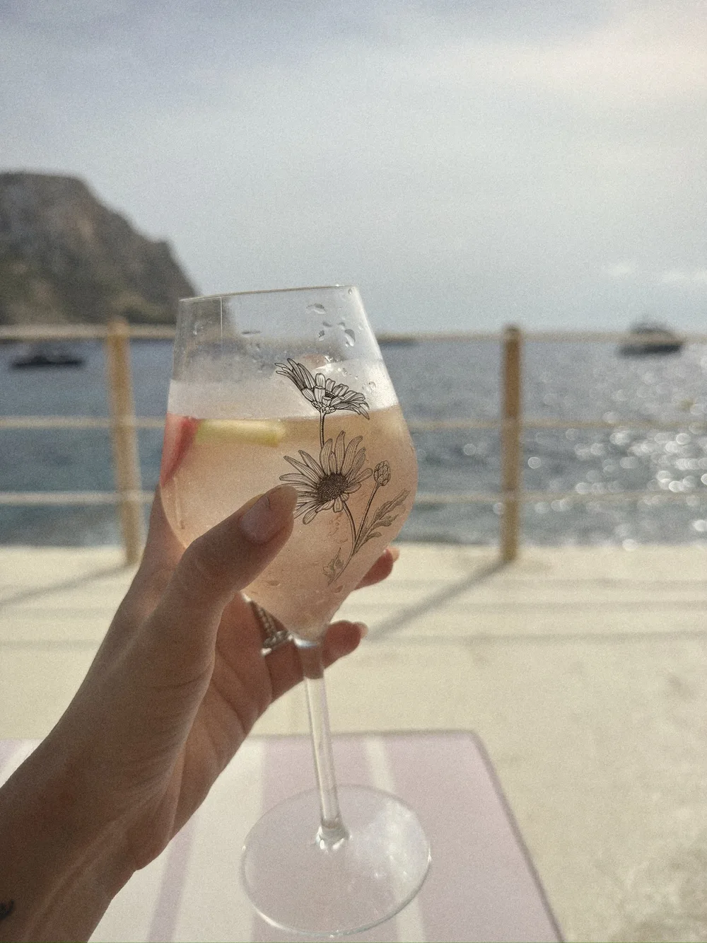 A hand holding a pale cocktail in a tall glass with a botanical print, the sea behind