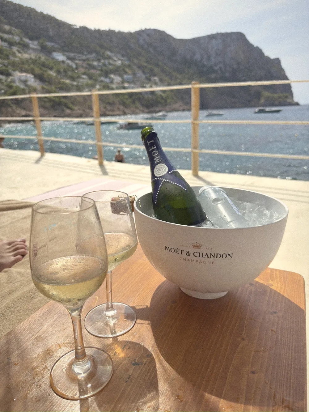 Two glasses of champagne and a bottle of Moët in an ice bucket on a wooden table, with the sea and cliffs behind