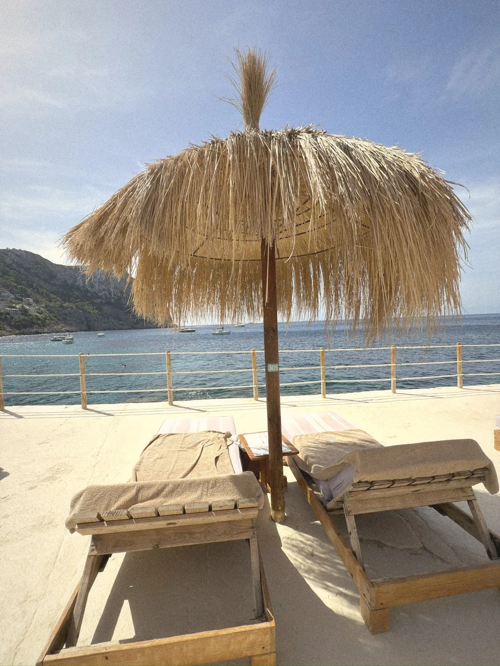 Two cushioned wooden loungers under a thatched straw umbrella, with the sea and mountains behind