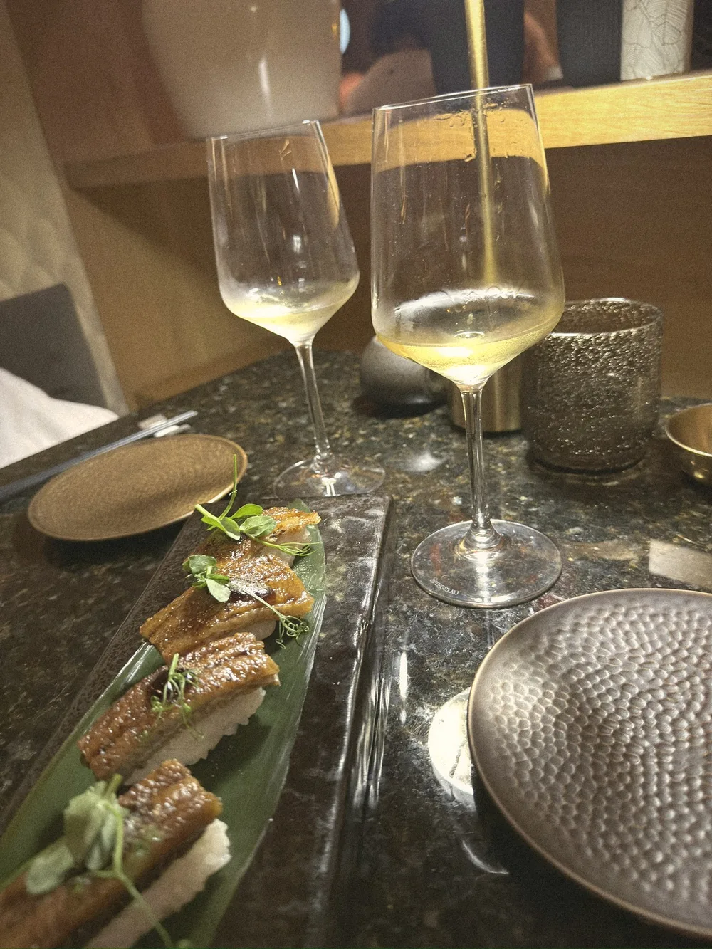 Glazed nigiri topped with microgreens on a dark plate, beside two glasses of white wine