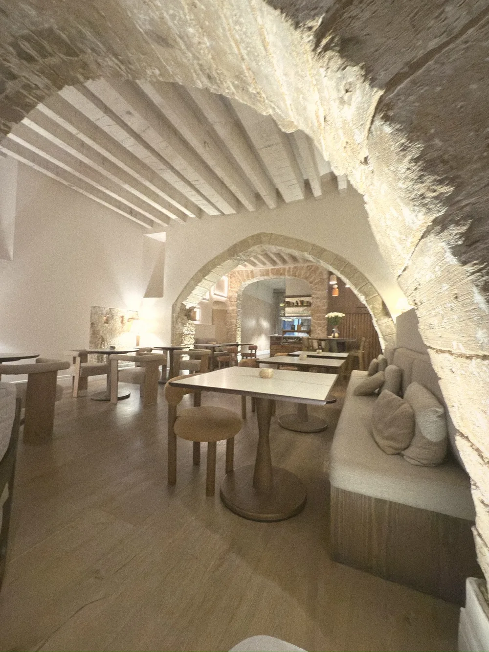 A softly lit dining room under whitewashed stone arches and wooden beams, with small tables and cushioned banquettes