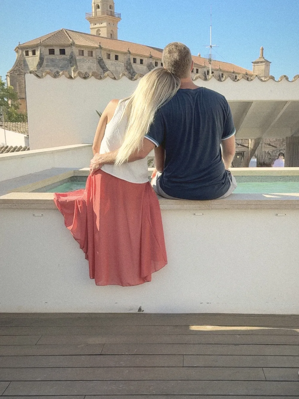 A couple seen from behind, sitting close together on the edge of the rooftop pool, looking at the basilica