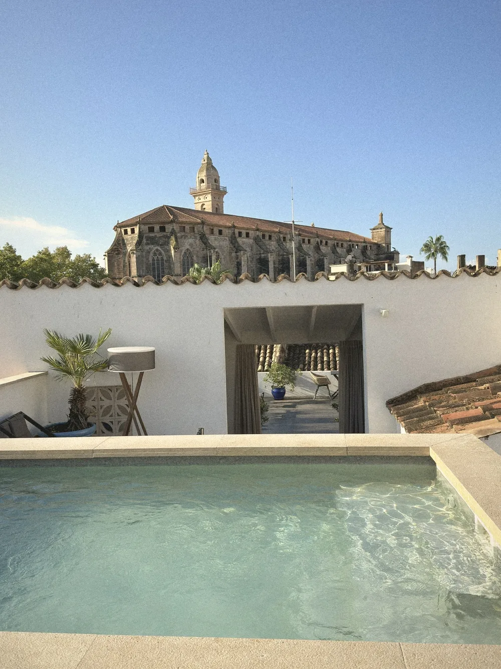 A small turquoise rooftop plunge pool behind a white wall, with the Gothic Basilica de Sant Francesc and its bell tower rising behind against a blue sky