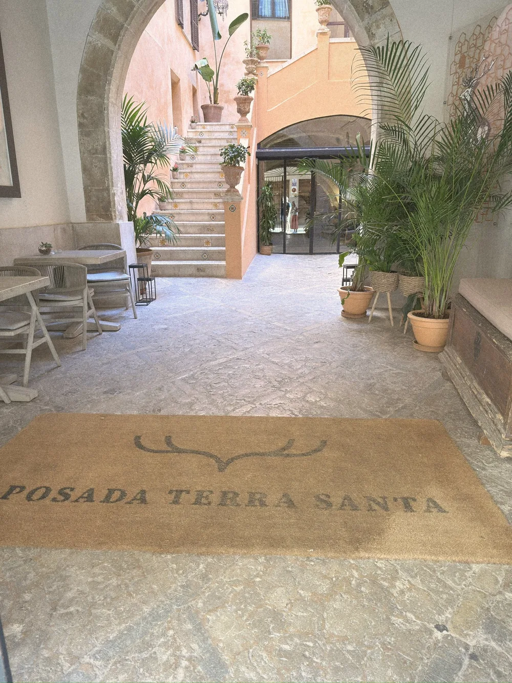 A doormat reading Posada Terra Santa at the entrance to a stone courtyard with palms and a staircase