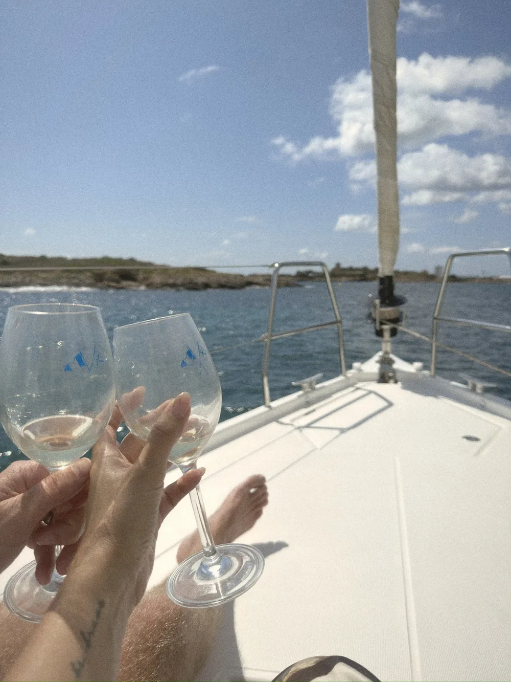 Two hands clinking glasses of white wine on the white bow of a sailing boat, with the blue sea and coast behind