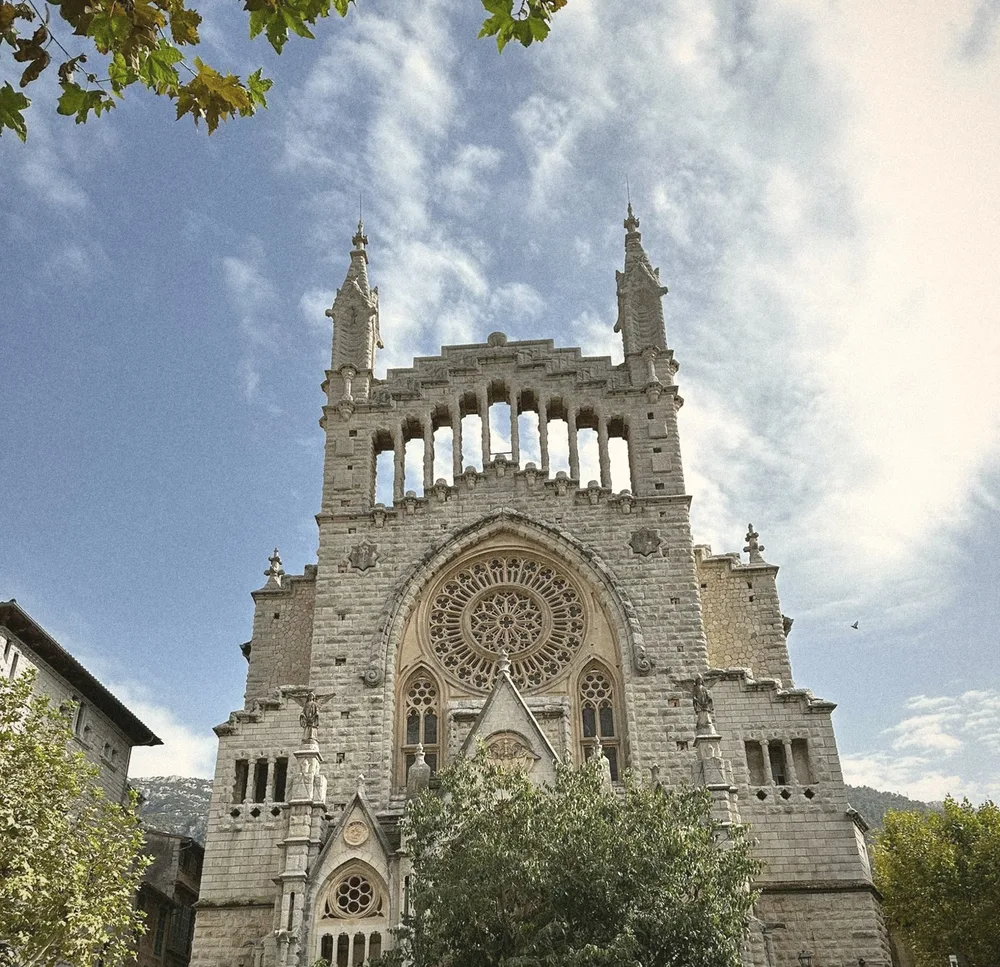 The ornate stone façade of the church of Sant Bartomeu in Sóller, with a rose window and twin spires against a blue sky