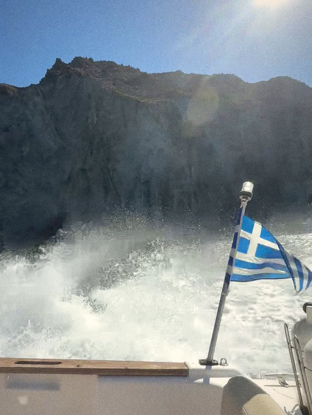 The Greek flag flying at the stern of a small boat, with white wake and dark cliffs behind