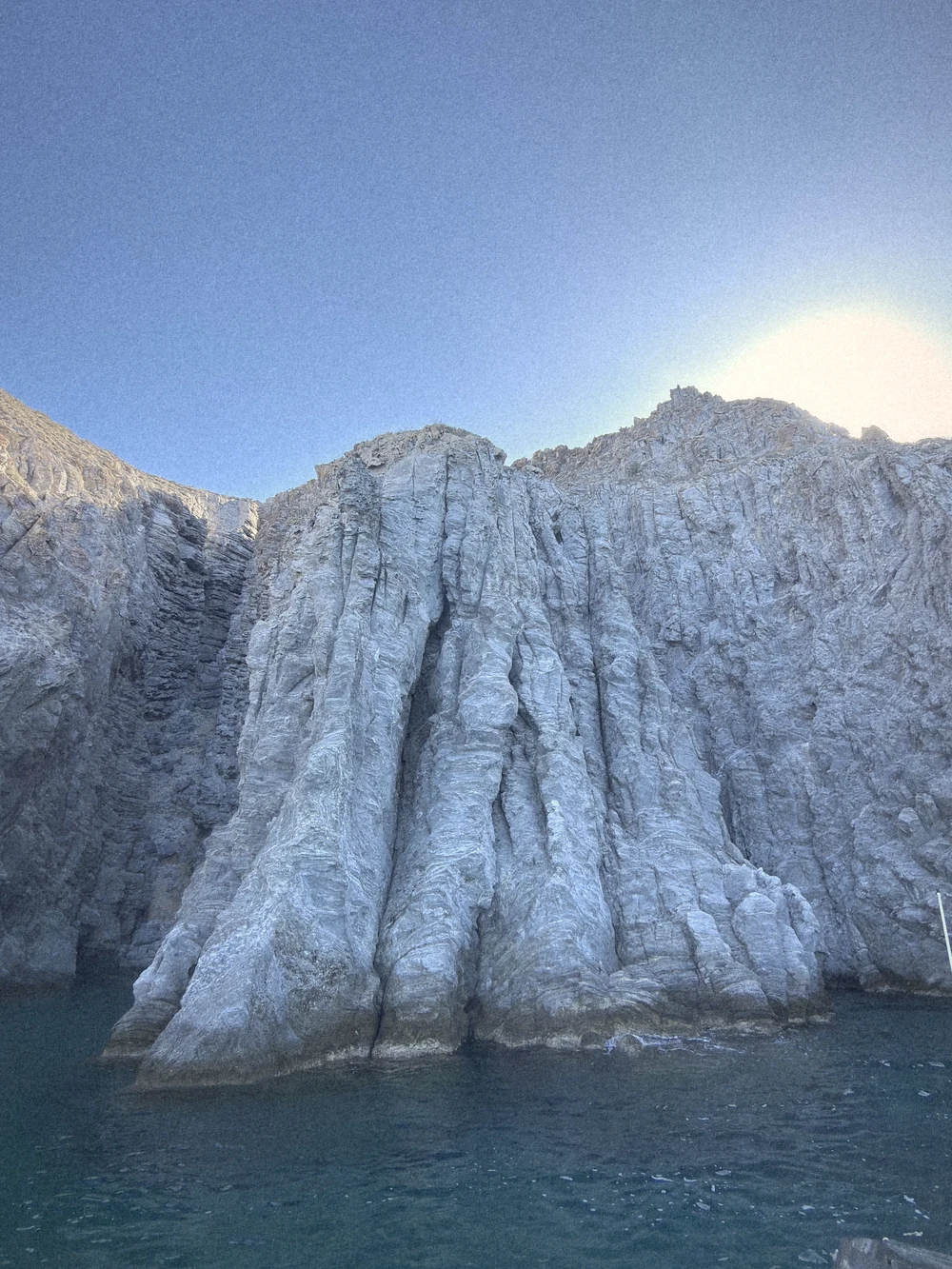 Tall grey ribbed rock cliffs rising out of the sea under a bright sky