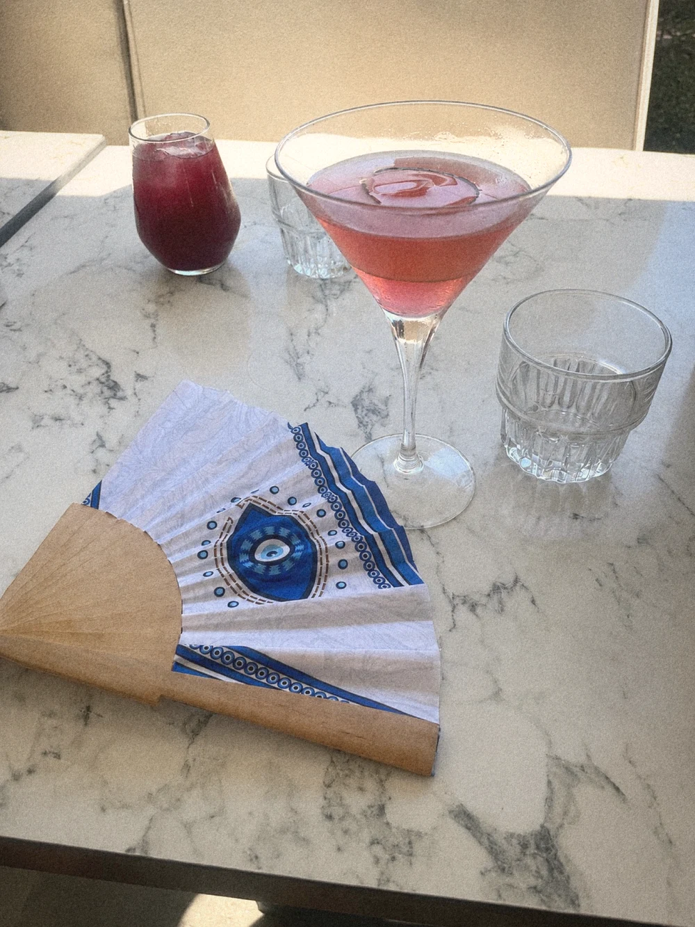 A pink cocktail in a coupe beside an evil-eye hand fan on a marble table