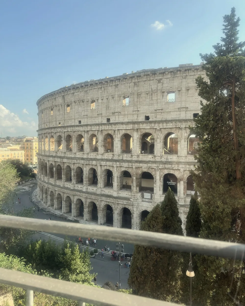 The Colosseum seen from a balcony between tall cypress trees under a blue Roman sky