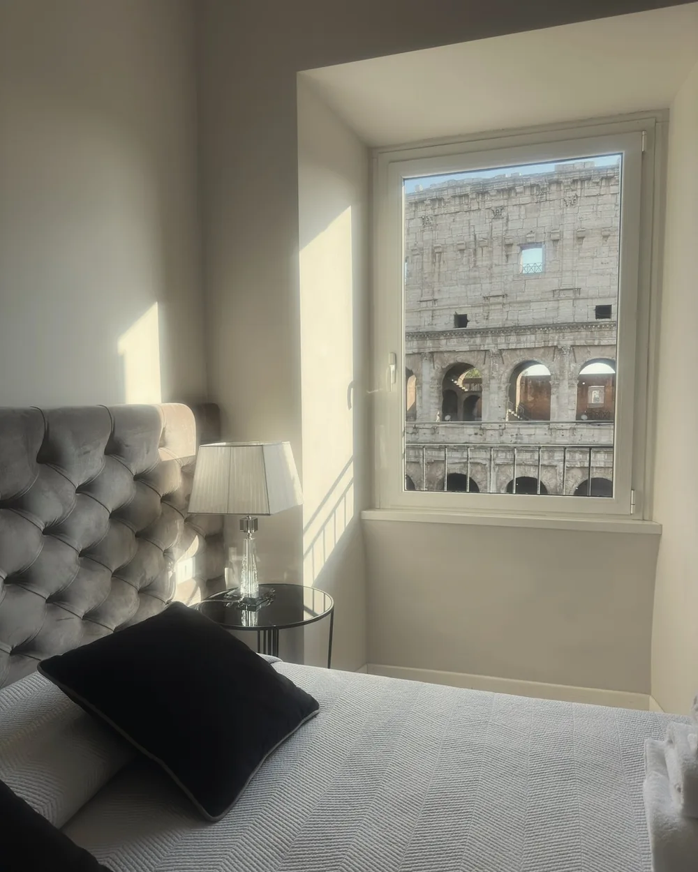 A soft white bedroom with a tufted headboard and the Colosseum filling the tall window